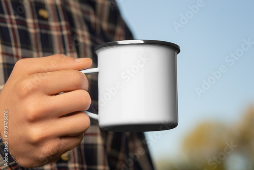 Mockup of a white enamel mug with a rim on a hand in a flannel shirt on a sky background, a metal cup for coffee, tea.