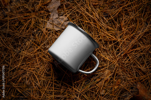 Mockup of enameled white mug with iron rim, handle, rustic cup for coffee, tea, on a background of pine needles, nature, outdoors.