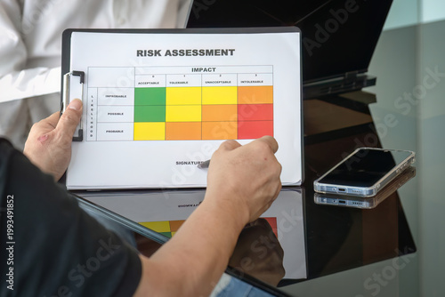 Over the shoulder shot of businessman holding pen over risk assessment report with colored matrix grid during office meeting.