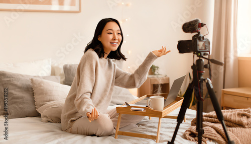 A woman sits on a bed, smiling and speaking to a camera on a tripod, with a laptop and coffee on a small table beside her in a cozy, light-filled room.