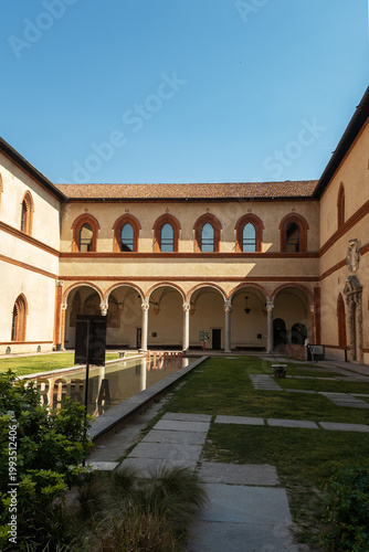 ​​A tranquil courtyard within the historic Castello Sforzesco in Milan, featuring elegant brick arches, a reflecting pool, and lush green lawns under a clear sky.


