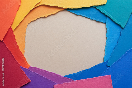 Overhead flat lay of rainbow-colored torn paper sheets arranged in a circular frame around a blank beige center, creating copy space for text or design use.