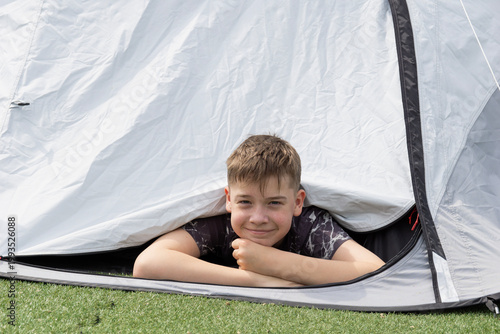 Fotografía a smiling teenage boy peeks out from a gray camping tent while lying on his stom