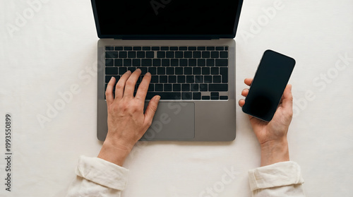 Overhead view of hands typing on laptop and holding smartphone, modern workspace setup