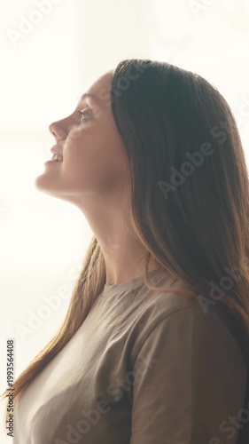 A woman gazes upward with a thoughtful expression. The woman has long hair and is illuminated by natural light, enhancing her contemplative demeanor.