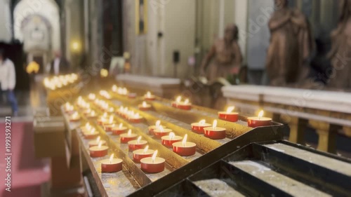Rows of red tea light votive candles burning with a warm glow on a metal stand inside a church, creating a serene and spiritual atmosphere for prayer, remembrance, and devotion