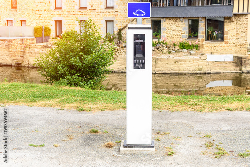 Charging station for electric cars in a deserted gravel car park along a river on a sunny summer day. A stone residential building is in background. 