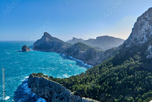 The rugged and wild coastline of northern Mallorca as seen from a high altitude, showcasing the dense pine forests and dramatic geological formations of the area.