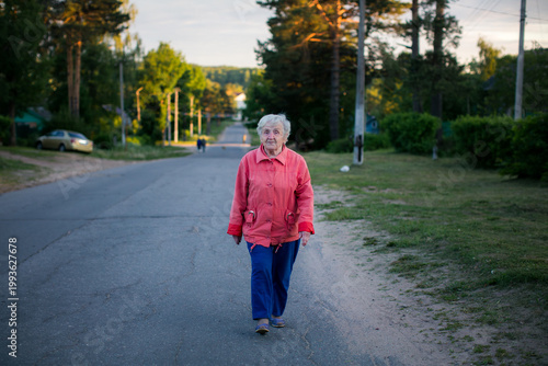 Elderly woman walking alone along a quiet rural road at sunset, surrounded by trees and houses, everyday life scene with soft evening light.