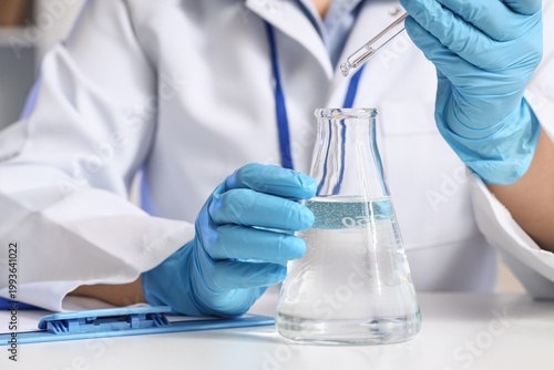 Female scientist dropping water sample into flask at table in laboratory, clo...