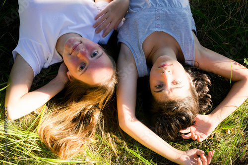 Close-up of two girls lying on grass, looking at the camera, warm sunlight on their faces, relaxed summer moment outdoors.
