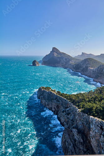 The vibrant turquoise waters of the Mediterranean Sea crash against the steep vertical cliffs of the northern Mallorca coast on a bright sunny day.