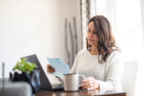 Senior woman at home kitchen table feeling stressed and confused while looking at multiple bills working on her laptop