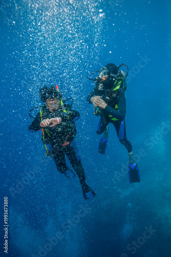 Two scuba divers hovering in blue water with ascending air bubbles, one checking his dive computer.