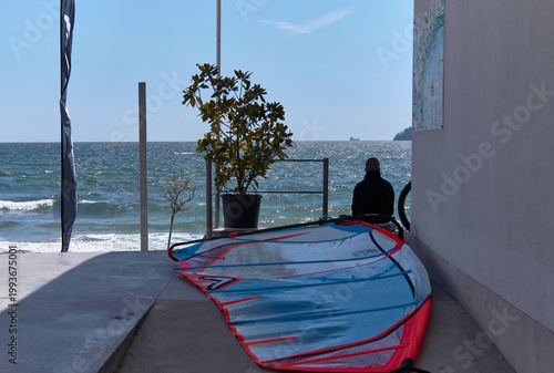Tableau sur toile Windsurfing equipment rests on a concrete surface while a person sits nearby, ov