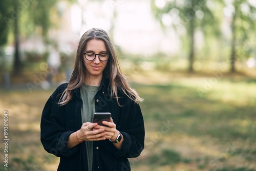 Young woman with glasses reading a message on her phone while standing in the park. High quality photo