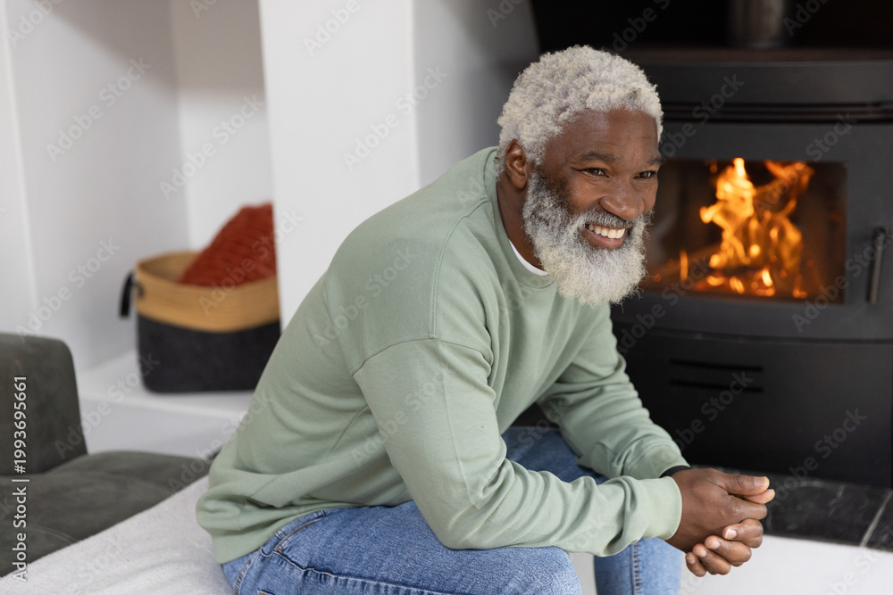 Obraz premium Senior African American man wearing green sweatshirt sitting in living room by wood stove, smiling