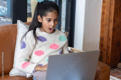 Asian child reacting with surprise on couch using silver laptop at home near dark-glass doors