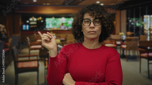 Woman wearing glasses crossing her arms beside empty chairs in a restaurant building entrance overlooking a bar counter; defiance.