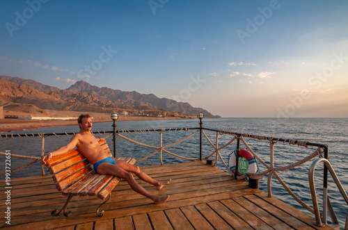 Man relaxing on a wooden pier at sunset with desert mountains and Red Sea in the background, Dahab, Egypt.
