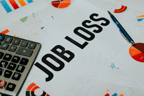 A document displaying the words Job Loss is placed on a table with graphs and charts surrounding it. A calculator and a pen are also visible, suggesting a financial assessment of employment changes