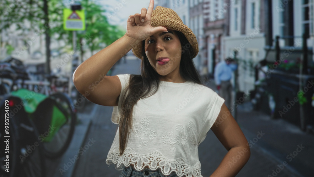 Fototapeta premium Young hispanic woman forms l with hand on forehead while sticking out tongue, wearing straw hat and white lace top on a city street; playful mischief.