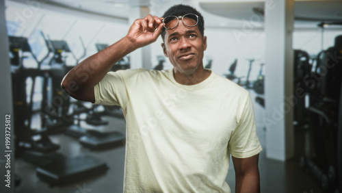 Fotografija Man lifting glasses with right hand, squinting while inspecting gym equipment in a fitness building; confusion