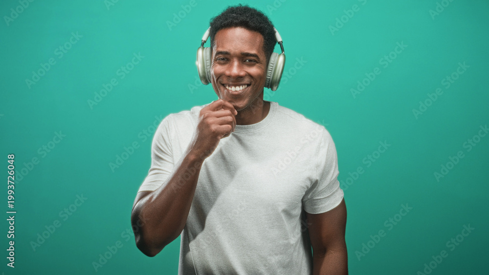 Naklejka premium African american man wearing over ear headphones, smiling with right hand to chin and relaxed posture against teal seamless studio backdrop; joyful confidence.