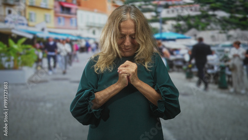 фотографія Senior woman clasping hands to chest, eyes closed in a prayerful gesture on a busy street with outdoor cafe umbrellas; quiet gratitude