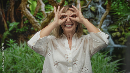 Фотография Caucasian middle age woman in a white blouse frames her eyes with hand binoculars in a lush forest setting; exploration joy