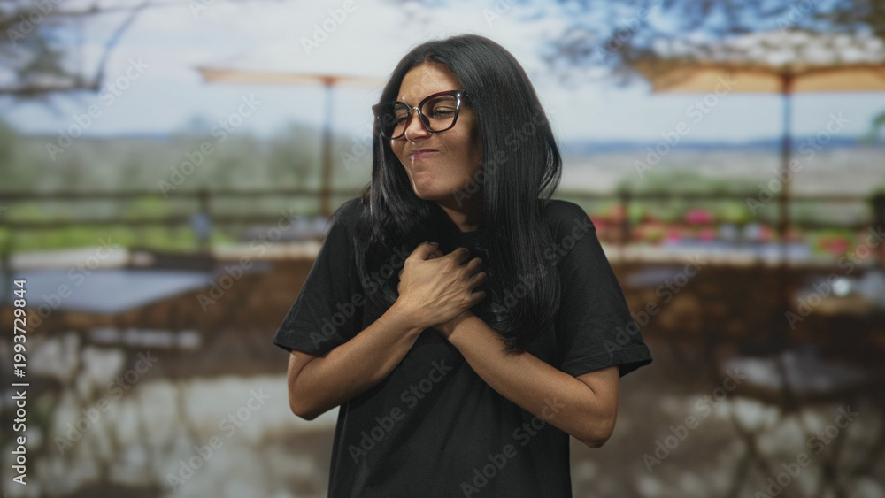 Fototapeta premium Woman with glasses and black shirt, hands clasped to chest with a wide smile showing teeth, standing on a building terrace with umbrellas and tables; joy gratitude warmth.