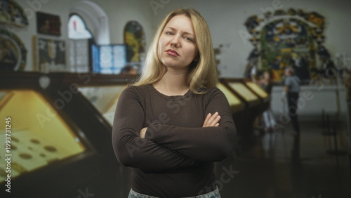 Принти на полотні Woman blonde with arms crossed pouting beside glass displaycases in a museum exhibition hall with other visitors nearby; frustration defiance isolation
