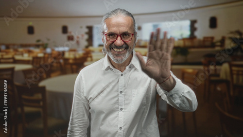 Canvas-taulu Man waves hand inside restaurant building, smiling and showing open palm while wearing white shirt and red glasses; friendliness welcome