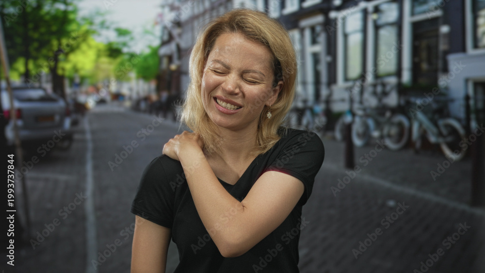 Fototapeta premium Blonde woman holds left shoulder on busy cobblestone city street lined with parked cars and bicycles under sunlight; discomfort.
