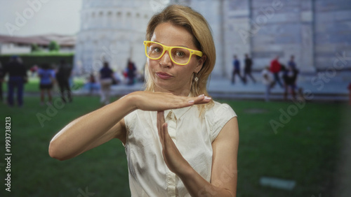 Blonde woman tourist hand forming timeout sign in front of the leaning tower of pisa building; reflection.