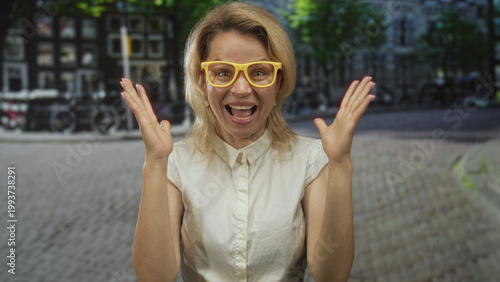 Canvas Print Blonde woman raises fists wearing yellow glasses and smiling on cobblestone street lined with bicycles; excitement