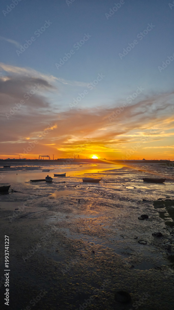 Fototapeta premium Vibrant sunset sky with dramatic clouds reflected on calm coastal water with small boats. Rich colors and contrast create a striking evening landscape.