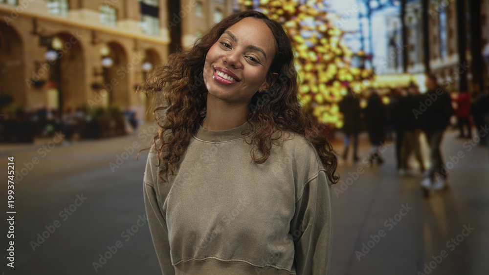 Fototapeta premium Woman smiles with visible face and slight head tilt on busy city street beside lit holiday tree and blurred crowd; joy confidence.