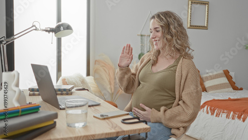 Obraz na płótnie Pregnant woman holding belly at laptop in bedroom, smiling and cradling bump beside bed and desk while seated; contentment