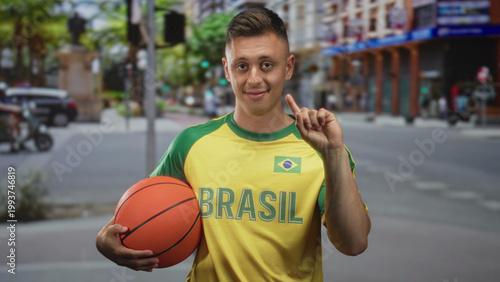 Young man holds a basketball with his hand and sometimes points finger, wearing a yellow brasil jersey in an urban street scene; confidence.