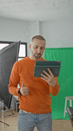 Fotografie Man holding tablet and gesturing with hand in studio among softbox lights and green screen, wearing orange sweater and jeans; creative concentration