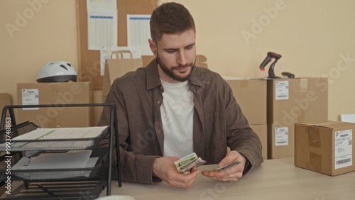 Картината върху платно Man counting dollars with hands at a packing desk surrounded by cardboard boxes, shipping labels and barcode scanner inside building; concentration diligence