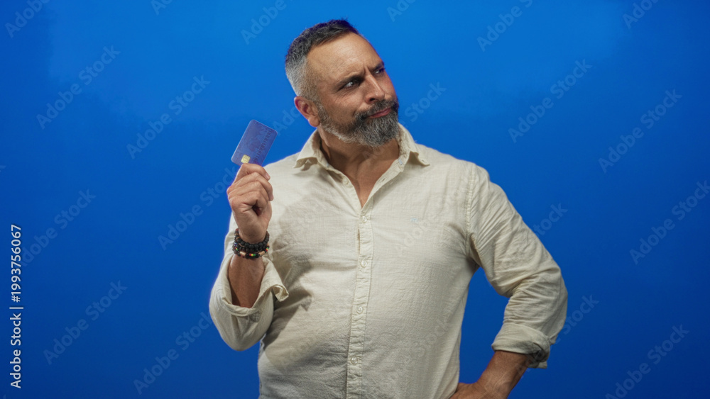 Fototapeta premium Man holding creditcard up with hand on hip and bracelets while posing in blue studio wall; confidence money choice.