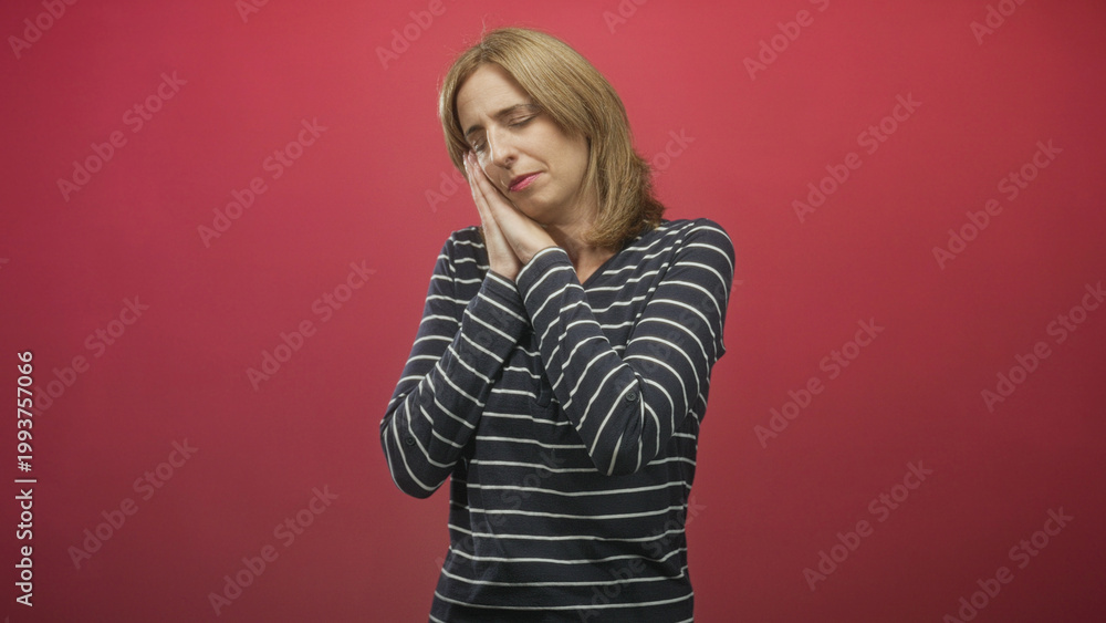 Fototapeta premium Woman with hands to cheek and closed eyes making sleeping gesture, wearing striped shirt against red studio backdrop; tired rest sleepiness.