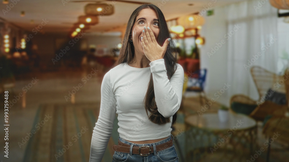 Fototapeta premium Young hispanic woman with hand over mouth smiling in a hotel lobby building wearing white top and jeans, casual pose; playful joy.