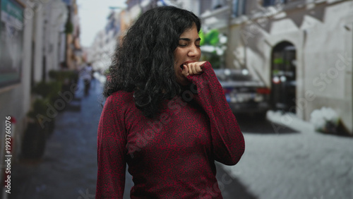 Картината върху платно Young brunette woman in red sweater bites her fist on a narrow city street, hand partly in mouth while suppressing a yawn and closing eyes; tired solitude