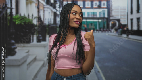 Принти на полотні Woman with exposed midriff in pink crop top smiling and pointing thumb to street, braided hair visible; confidence