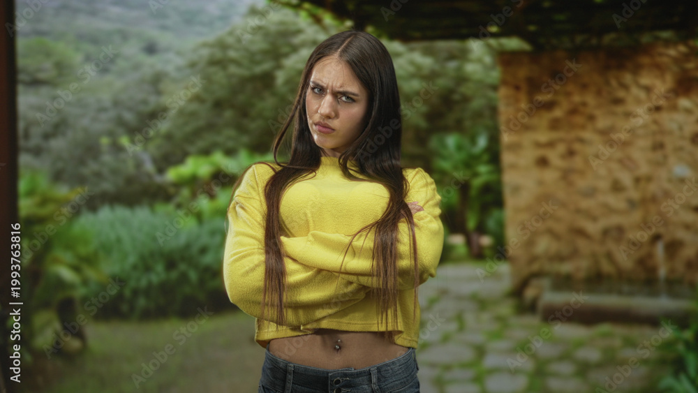 Naklejka premium Teenage woman arms crossed in a yellow sweater standing by a stone building with visible belly piercing; defiant attitude.