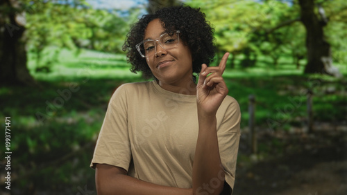 Photography Woman wearing glasses tilts head and points finger to object near lush green trees in park; confidence insight clarity