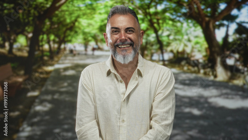 Man with gray beard smiling and folded arms on a tree lined street in a park under dappled sunlight; contentment.